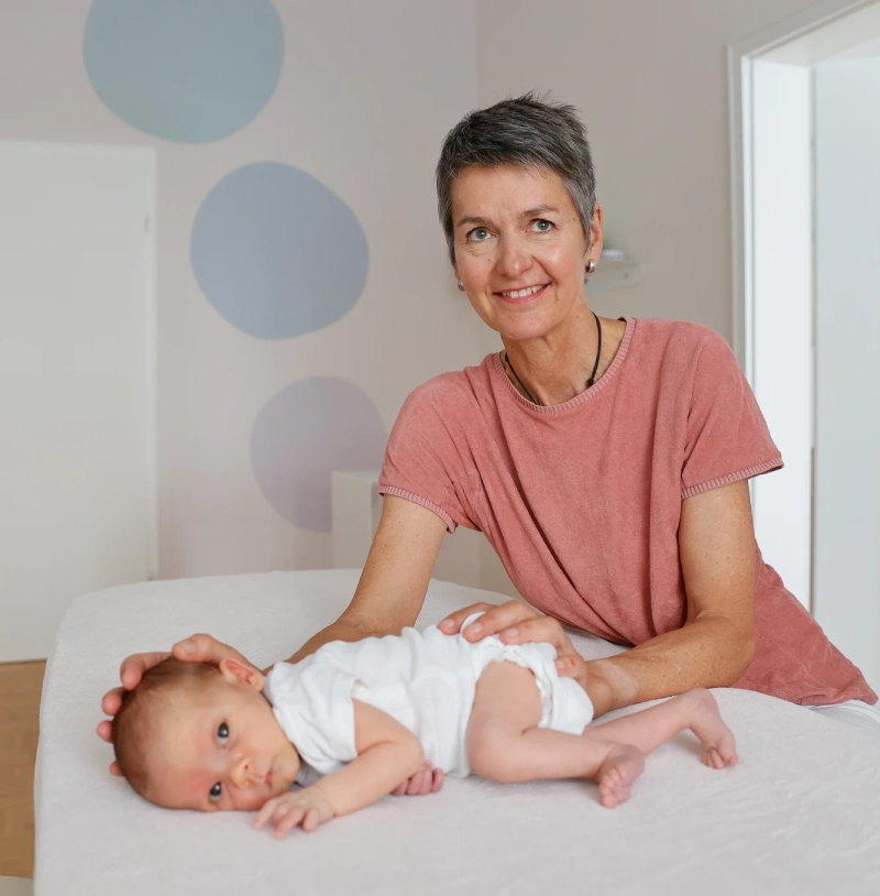 Eine lächelnde Frau in rosa Shirt hält ein Baby auf weißem Bettlaken mit pastellblauen Kreisen im Hintergrund