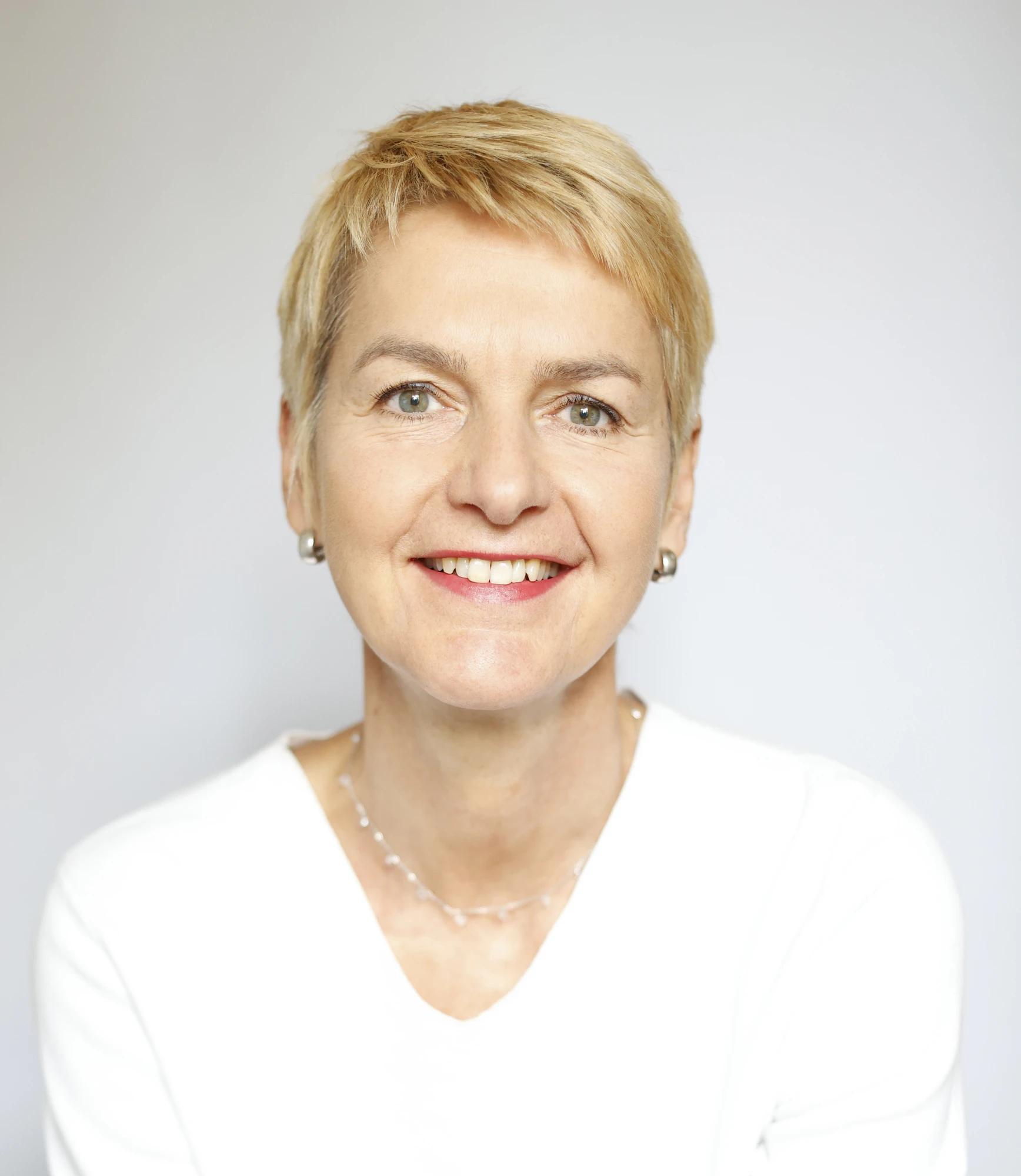 Lächelnde Person mit kurz geschnittenen blonden Haaren vor hellem Hintergrund in weißer Bluse mit Perlenkette und Ohrringen